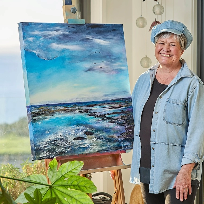 Marlyse standing by her easel with a seascape painting